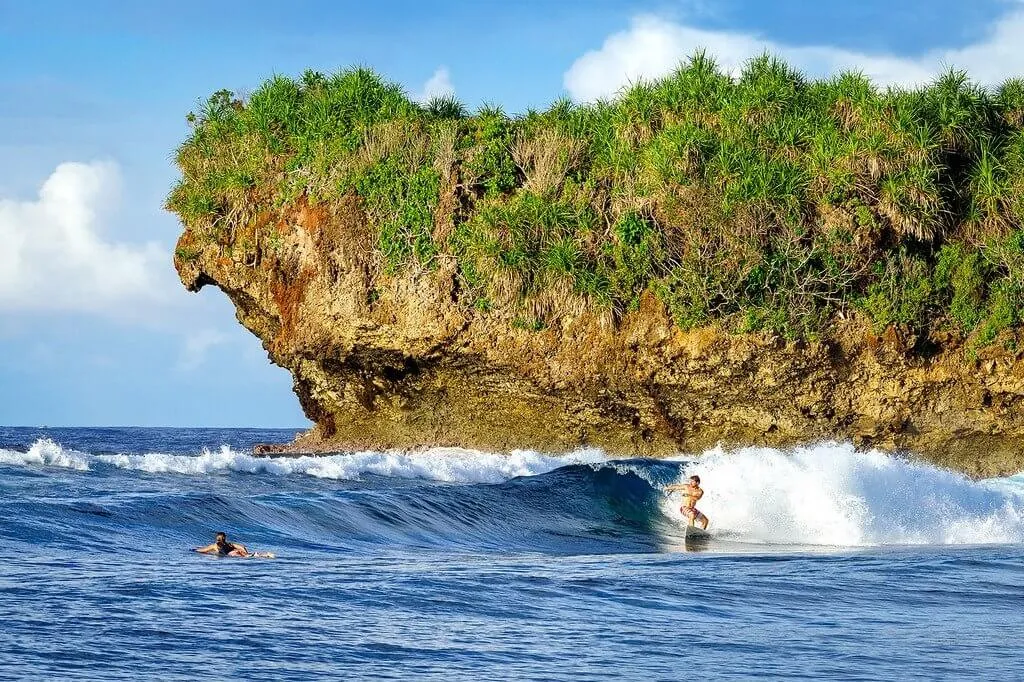 Surfers riding a wave beneath the jungle-topped cliffs of Rock Island near Siargao