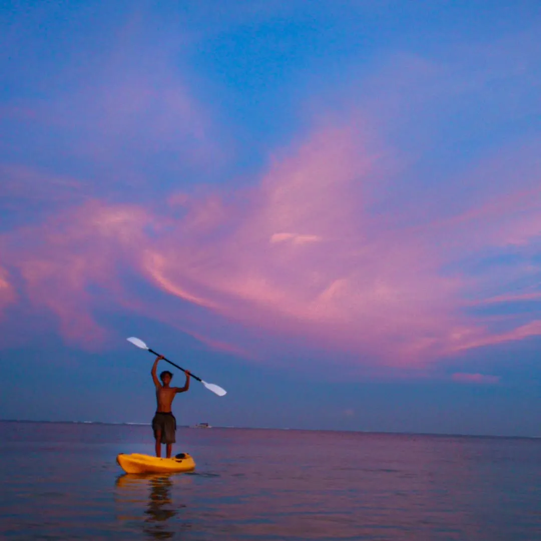 Paddler standing on a yellow kayak raising a paddle under a pink dusk sky in Siargao